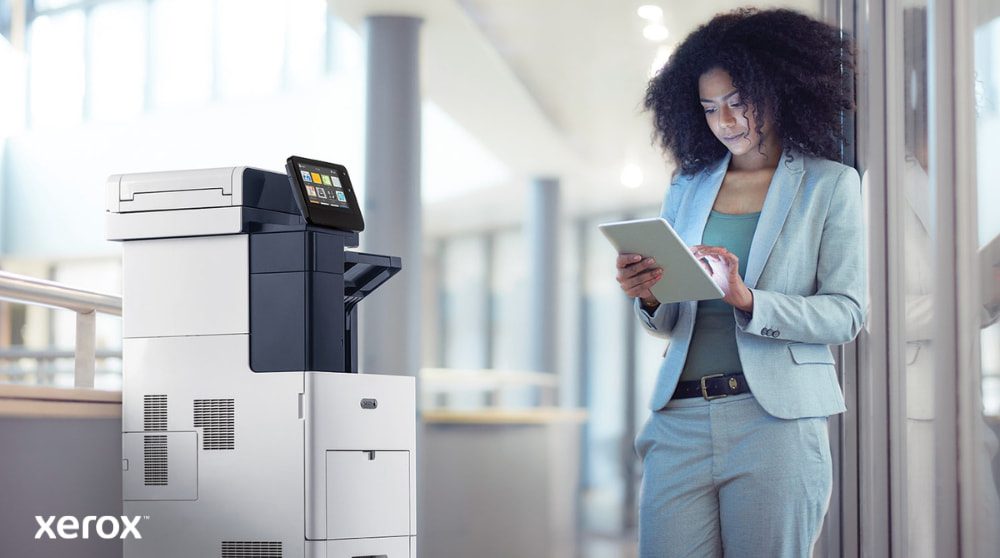 Xerox® ConnectKey® : connectivité mobile Une personne se tient à côté d'une imprimante multifonction Xerox VersaLink B605, regardant et interagissant avec une tablette, illustrant comment ConnectKey permet une interaction mobile avec l'imprimante.