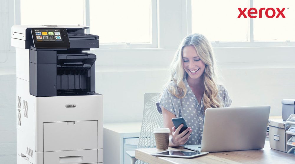 Xerox® ConnectKey®: Nahtlose Workflow-Integration Eine Person schaut zufrieden auf ihr Smartphone, während sie mit einem Laptop an einem Schreibtisch sitzt. Neben dem Schreibtisch in einem hell erleuchteten Büroraum ist ein Multifunktionsdrucker Xerox VersaLink B605 zu sehen.