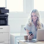 Une personne regarde joyeusement son smartphone, assise à un bureau avec un ordinateur portable. Une imprimante multifonction Xerox VersaLink B605 est visible à côté du bureau, dans un espace de travail lumineux.