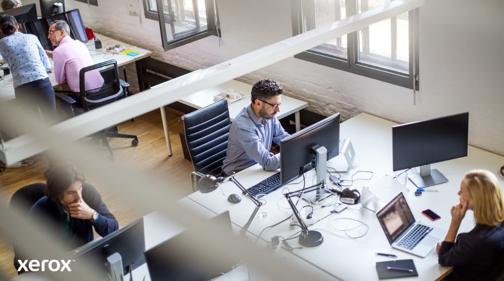 Eine Draufsicht auf einen Großraumbüroraum mit mehreren Personen, die an ihren Schreibtischen am Computer arbeiten. Die Szene vermittelt den Eindruck einer produktiven und kollaborativen Arbeitsumgebung.