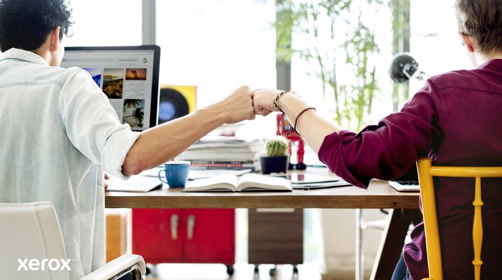 Xerox® Smart Start: Teamarbeit und Zusammenarbeit Zwei Personen sitzen an einem Schreibtisch und blicken von der Kamera ab. Sie stoßen sich mit den Fäusten aneinander, um Teamwork und erfolgreiche Zusammenarbeit zu symbolisieren. Eine Person blickt auf einen Computerbildschirm.